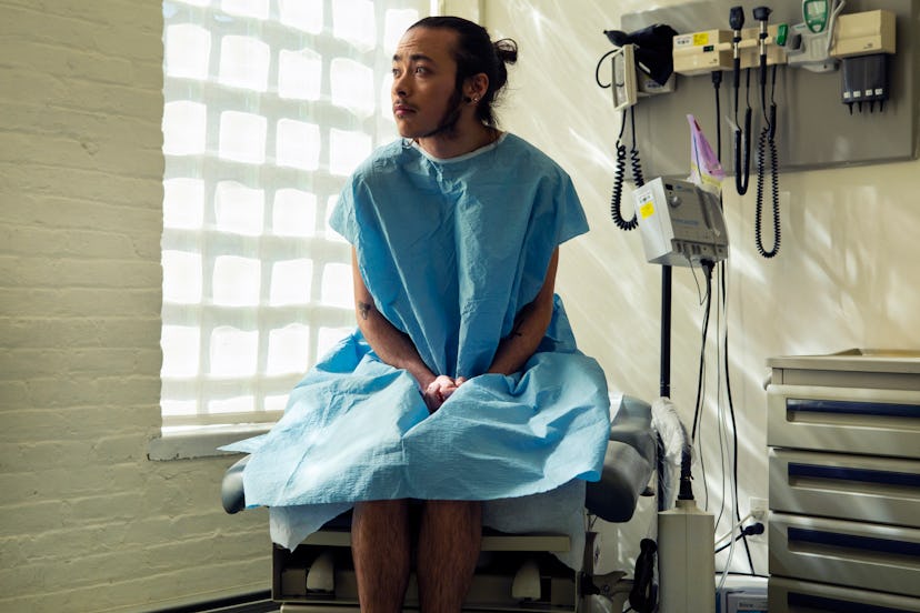 A genderqueer person in a hospital gown sitting in an exam room at a doctor's office.