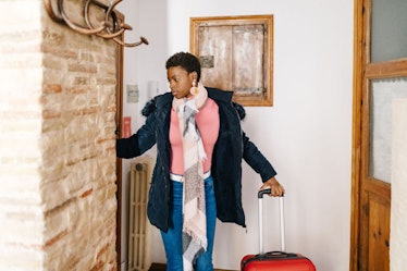 A woman wearing a jacket, sweater, scarf, and jeans stands near the door of a cozy apartment with a ...