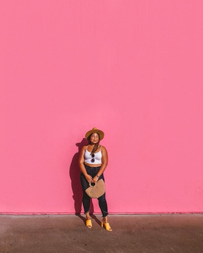 A woman in a white tank top, blue jeans, and yellow heels standing in front of the Paul Smith pink w...