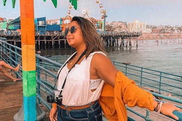 A woman in sunglasses, an orange sweater, tank top, and jeans hangs out at Santa Monica Pier on a su...