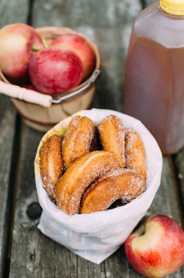 A bag of apple cider doughnuts sits on a picnic table at a cider mill next to a basket of apples and...
