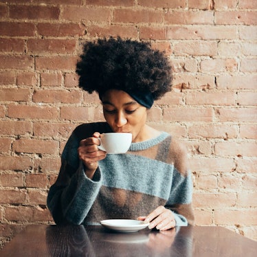 A black woman sips on hot cider in a mug in front of a brick wall at a cider mill.