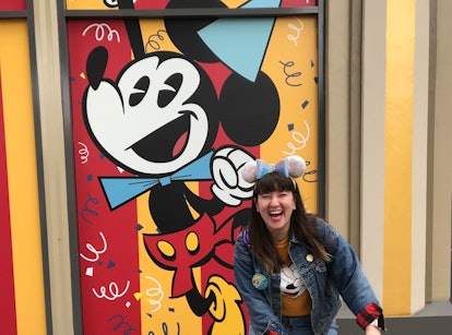 A woman wearing sequinned Minnie ears and a Disney birthday button laughs in front of a Mickey Mouse...