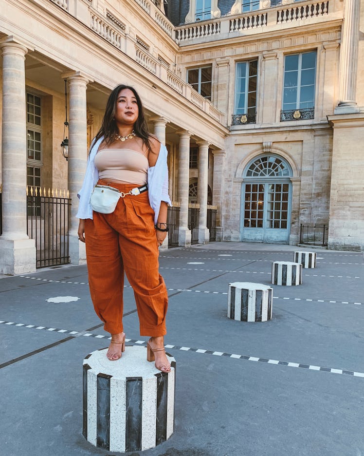 A woman in a flowy white button-up, nude crop top, and baggy orange pants stands on top of a black a...