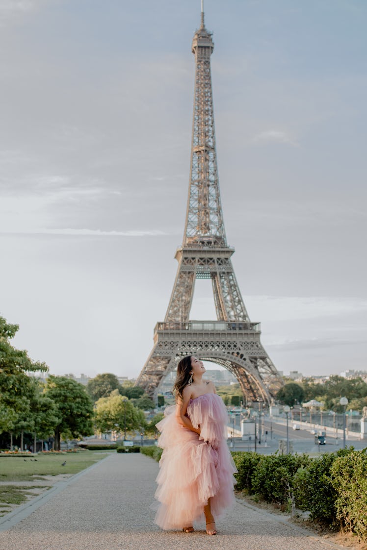 A girl dressed in a pink tulle dress smiles, looks up, and poses in front of the Eiffel Tower in Par...