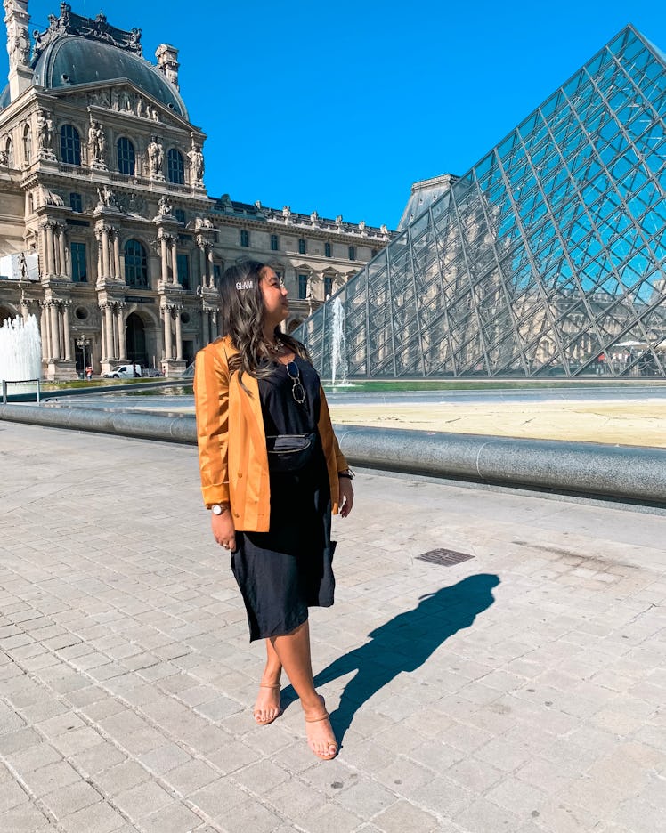 A woman dressed in a black slip dress and bright yellow-gold blazer poses in front of the Louvre Pyr...