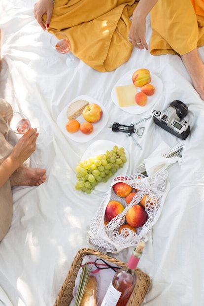 A picnic blanket is on the grass with grapes, wine, bread, fruit, and a film camera for the ultimate...