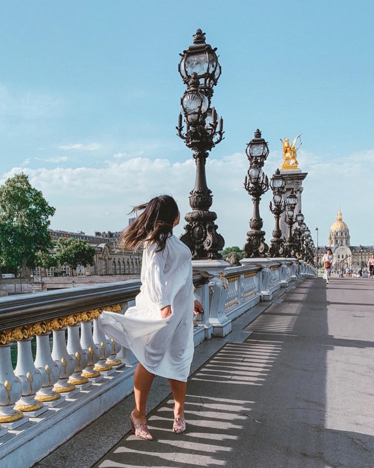 A woman in a flowy white dress poses with her hair blowing in the wind on the Pont Alexandre III. 