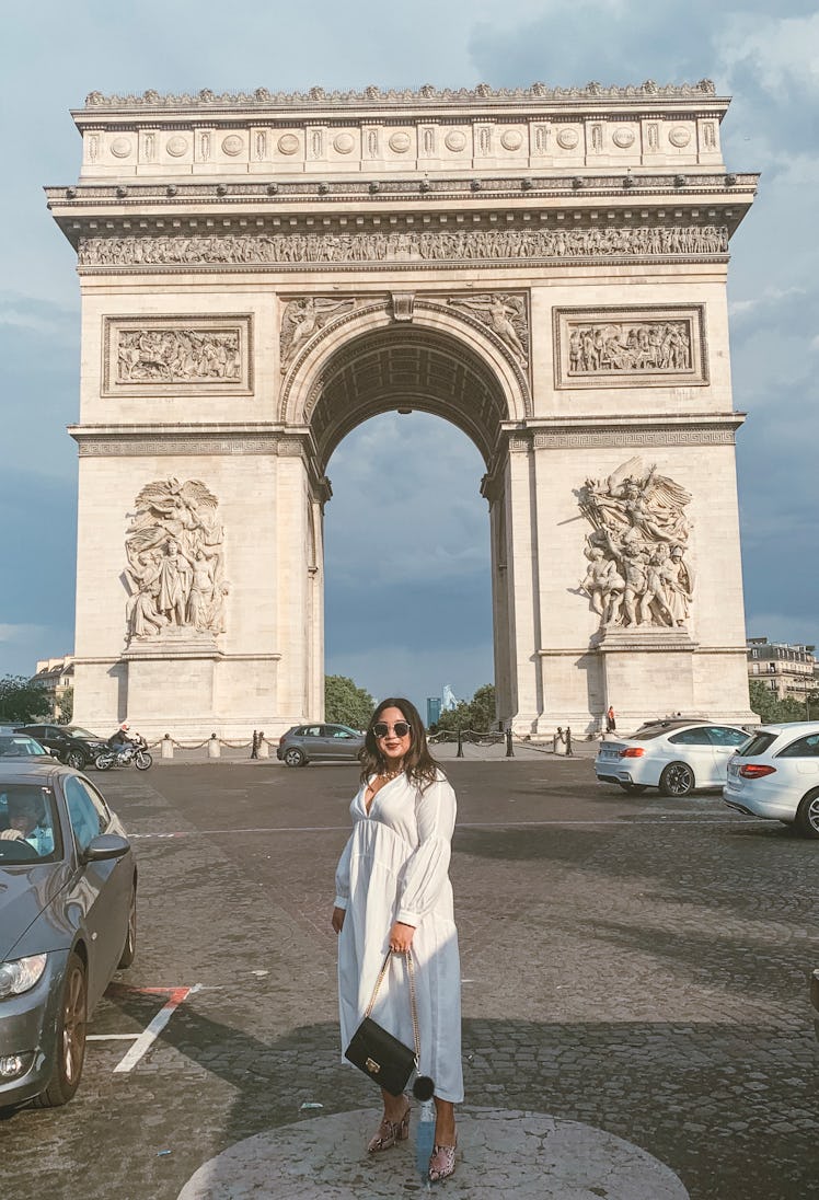 Woman in a flowy white dress holding a black purse on a chain standing in front of the Arc de Triomp...