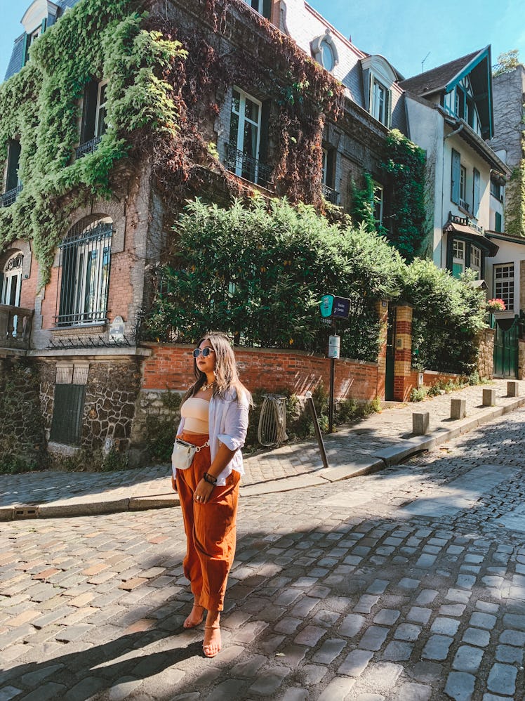 A woman in a flowy white button-up, nude crop top, and baggy orange pants stands on a cobblestone st...