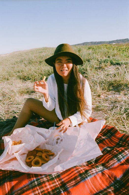 A woman eats fish and chips on a red plaid picnic blanket on a green hill in the fall.