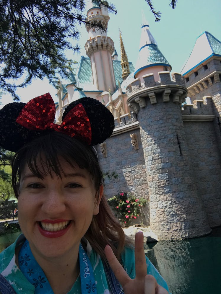 A woman smiles, holds up a peace sign, and takes a selfie in front of the castle at Disneyland with ...