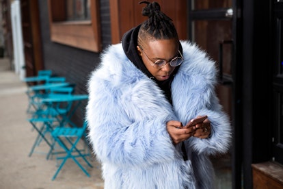 A transmasculine person with a furry blue coat checking his phone on the sidewalk. Your brain likes ...