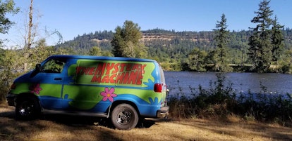 The Mystery Machine from 'Scooby Doo' sits near a lake, surrounded by a bunch of trees.