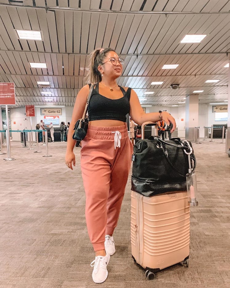 A woman in black crop top and pink sweatpants with carry-on luggage waits at an airport gate.