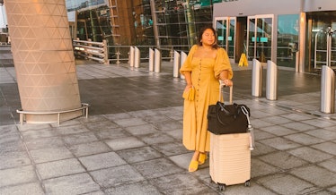A woman in a long button-down yellow dress and yellow shoes stands in front of an airport with her b...