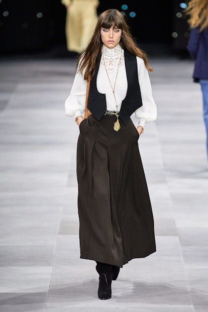 a model going down the runway wearing a white button-up shirt, a long black skirt and a waistcoat