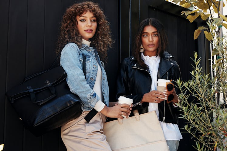 Two girls stand in front of a black fence with coffee cups and bags from Shay Mitchell's BÉIS winter...