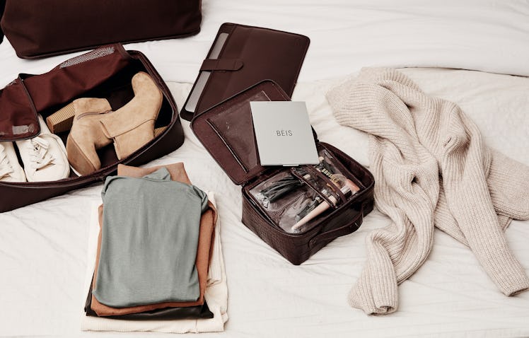 Winter clothes, shoes, and beauty products are laid out on a white bed and packed into espresso-colo...