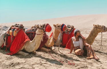 A woman in a white tank top and brown pants with a scarf wrapped around her head sits in the sand su...