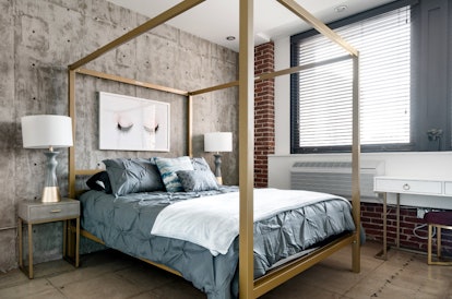 A queen bed in an industrial-looking room covered in slate gray bedding surrounded by a gold canopy ...