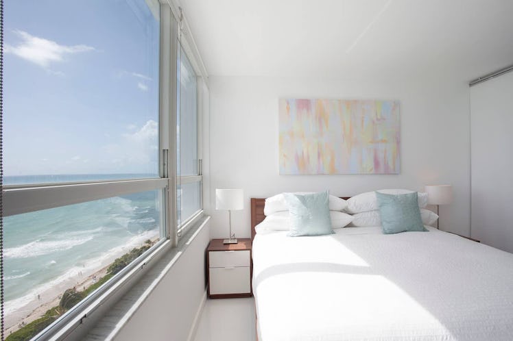 A room with a white queen-sized bed and wooden bedside tables overlooks the beach and an ocean. 
