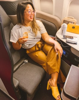 A woman wearing a white T-shirt, brown pants, and yellow mule heels sits in a business class plane c...