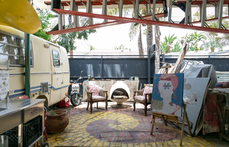 An outdoor art studio with murals and paintings scattered around and a vintage camper on the left. 