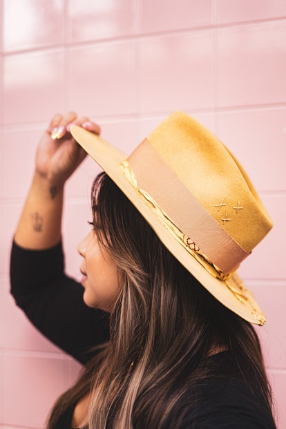 A profile of a woman with dark hair wearing a mustard yellow felt hat against a pink tile wall. 