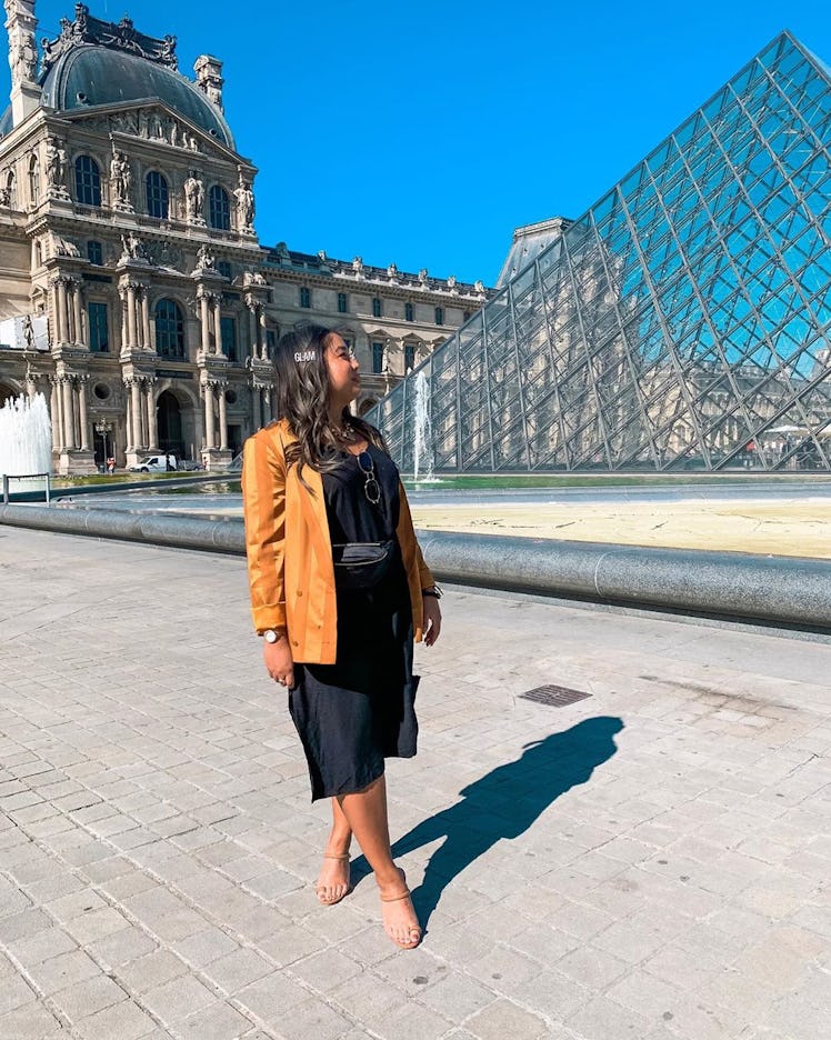 A woman in a black dress and yellow blazer poses in front of the Louvre Pyramid with her head turned...
