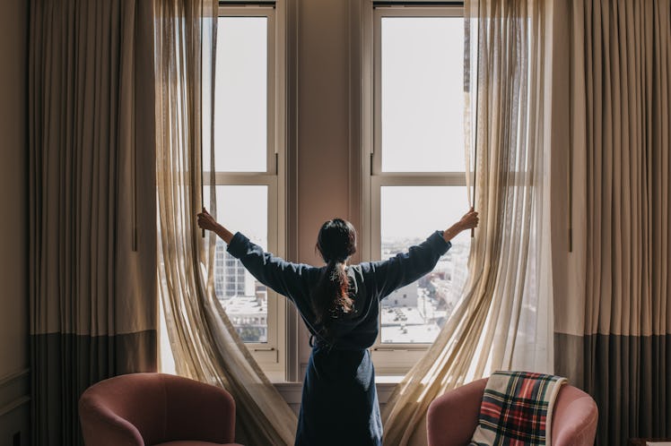 A woman in a bathrobe with her back turned to the camera opens up the curtains in her room.