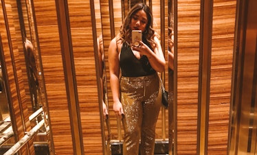 A woman in a black halter top and sparkly gold pants takes a phone selfie in an elevator mirror.