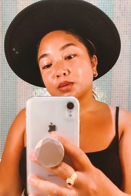 A woman in a black tank top and black hat takes a closeup phone selfie in an elevator mirror.