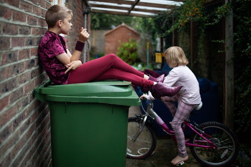 A transgender girl sits on a garbage bin in an alley with her sister on a bicycle.