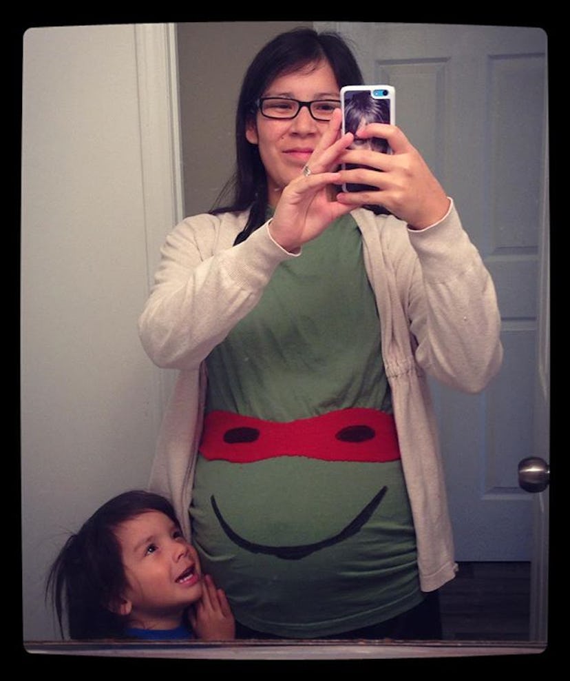 A pregnant woman wearing a ninja turtle tank top taking a mirror selfie with her kid standing next t...