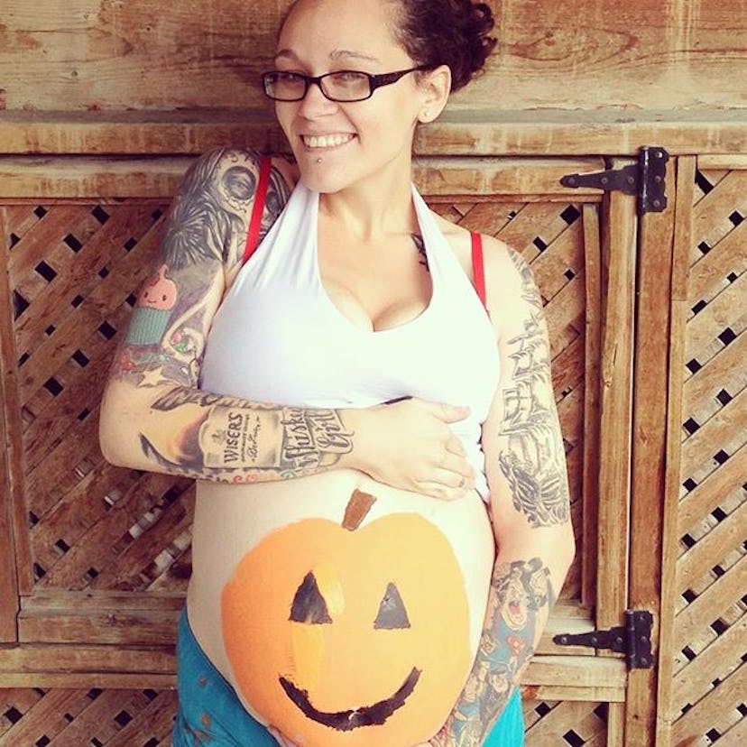 A pregnant woman posing for a photo, holding her belly with Jack O' Lantern painted on it.