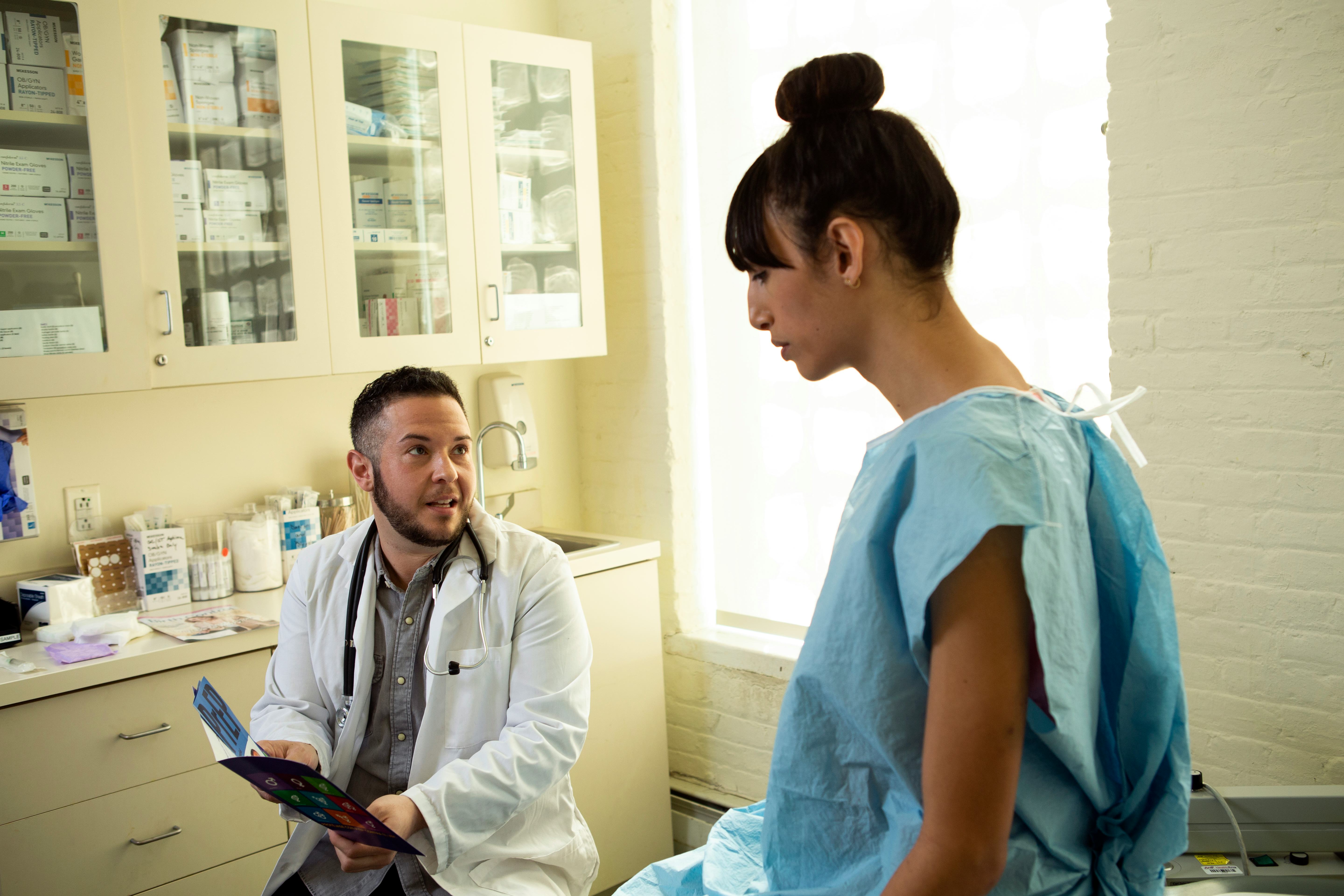 A transgender woman in a hospital gown speaking to her doctor, a transgender man, in an exam room. G...