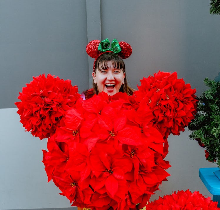 A woman stands behind a poinsettia flower bush shaped like Mickey Mouse, while wearing holiday Minni...