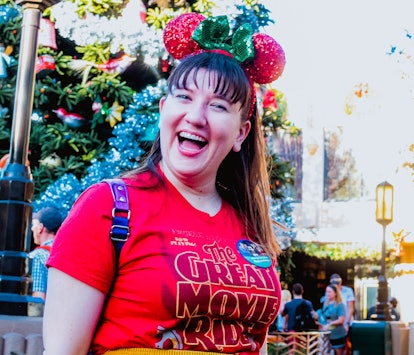 A woman wearing a holiday Disney button and festive Minnie ears smiles in front of a Christmas tree ...