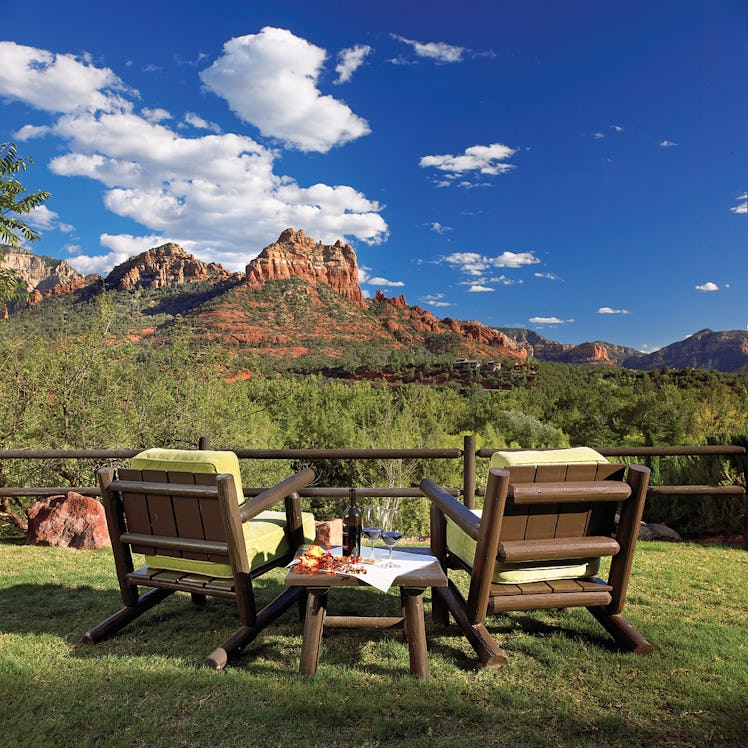 Two wooden lawn chairs and a matching table are on a lawn overlooking red rock mountains in the dist...