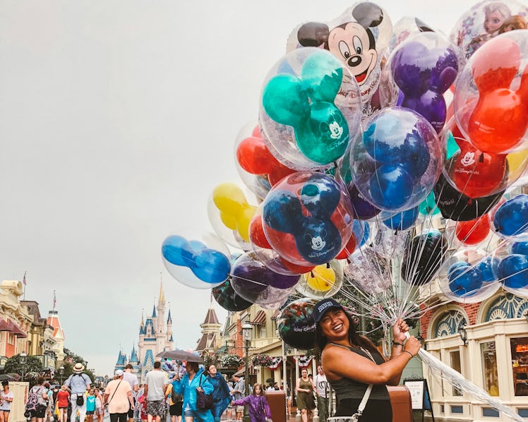 A woman wearing a baseball cap smiles and holds a bouquet of balloons in front of the Disney World c...