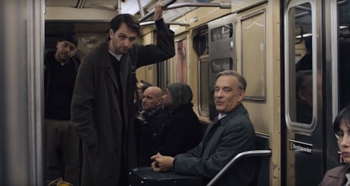 A dark-haired man in a long coat stands above an older man wearing a grey jacket and tie seated in a...