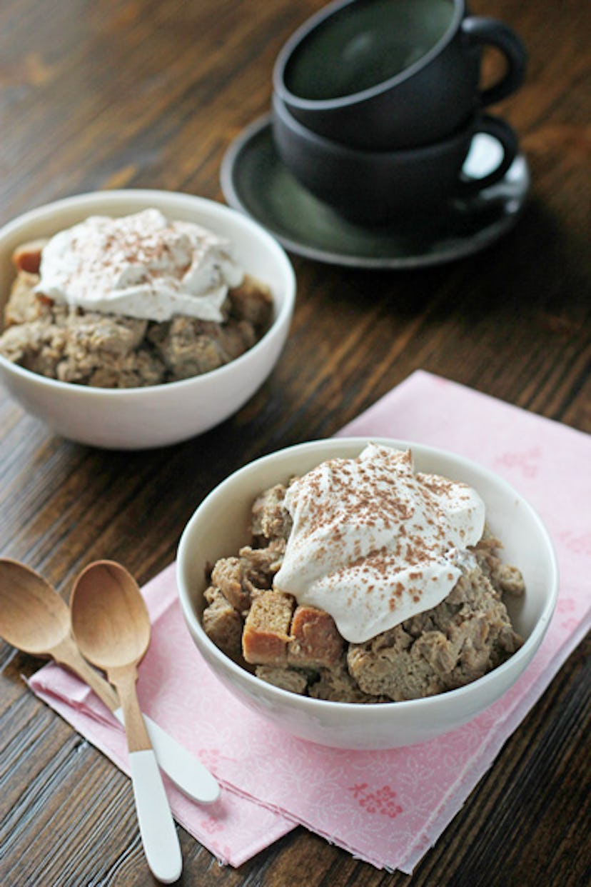 two bowls of bread pudding with cream on top placed next to two wooden spoons and stacked grey teacu...