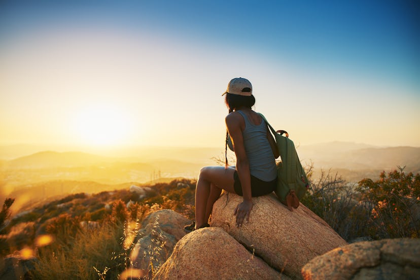 A person in a snapback and tank top with a green backpack sits on a rock while looking out over the ...