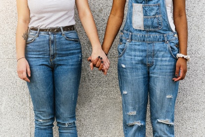 Two people wearing ripped jeans hold hands while standing in front of a concrete wall. When someone ...