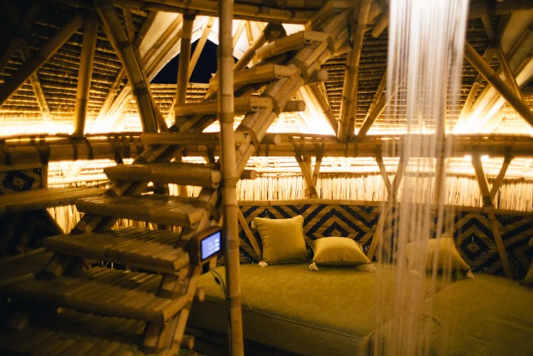 A dimly-liy room is surrounded by bamboo walls, with a bamboo staircase and a small couch. 