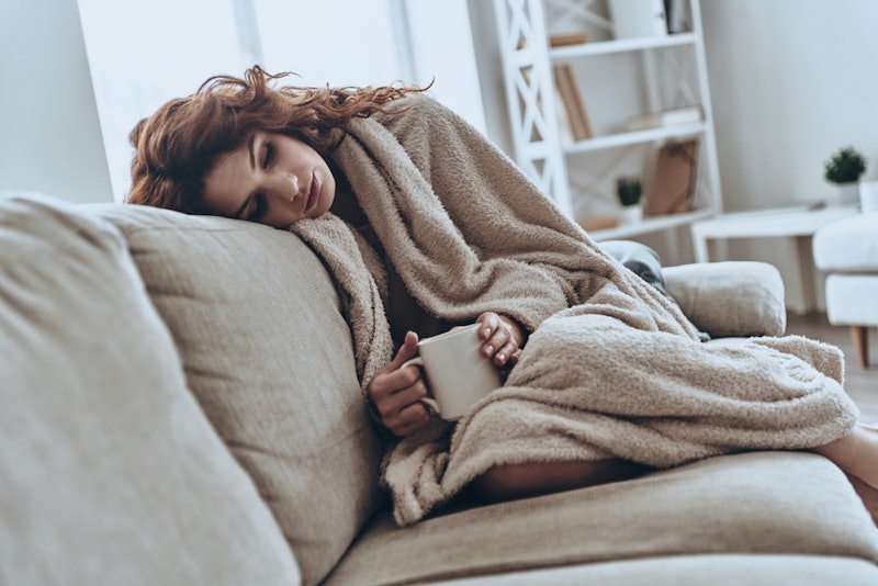 A person curls up on a couch with a blanket and a mug. Not getting enough sleep can make you more an...
