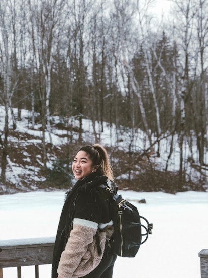 A smiling woman wearing a fuzzy coat and a backpack is surrounded by a field of snow and trees in th...
