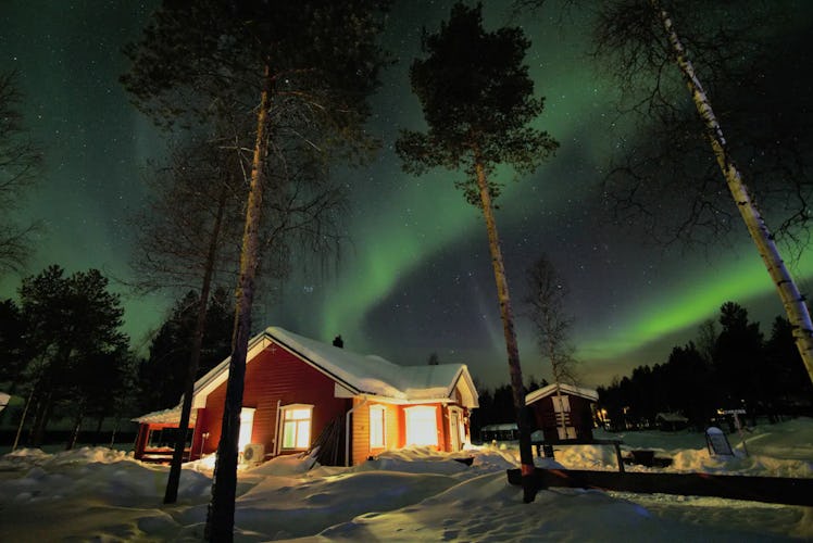 A cottage in the woods is covered in snow with the Northern Lights shining above. 