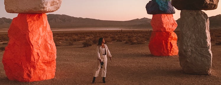 A woman stands in the desert surrounded by colorful rock installations looking off into the distance...